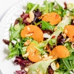A fresh mixed greens salad with sliced apricots, nuts, and garnished with green onions on a white plate.