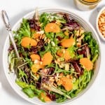 A fresh mixed greens salad topped with slices of mandarin oranges and chopped nuts, accompanied by a side of dressing.