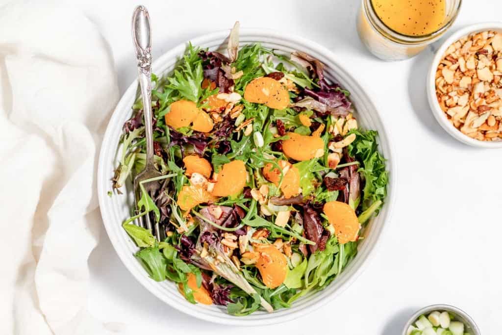A bowl of mixed greens salad with sliced oranges and chopped nuts, served with a dressing on the side.