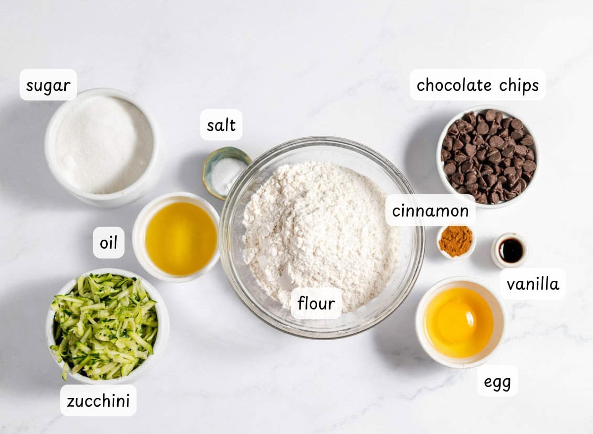 Baking ingredients for chocolate chip zucchini muffins&mdash;including flour, sugar, chocolate chips, zucchini, oil, salt, cinnamon, vanilla, and an egg&mdash;are arranged in bowls on a white surface.