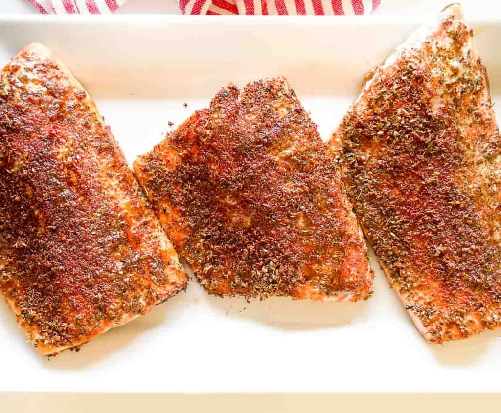 Three seasoned salmon fillets on a white plate, with a red and white striped napkin on the side.