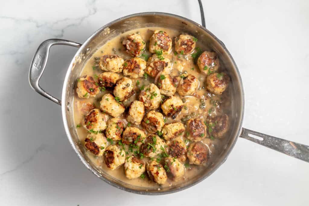 A skillet filled with cooked meatballs in a creamy sauce, garnished with chopped parsley.