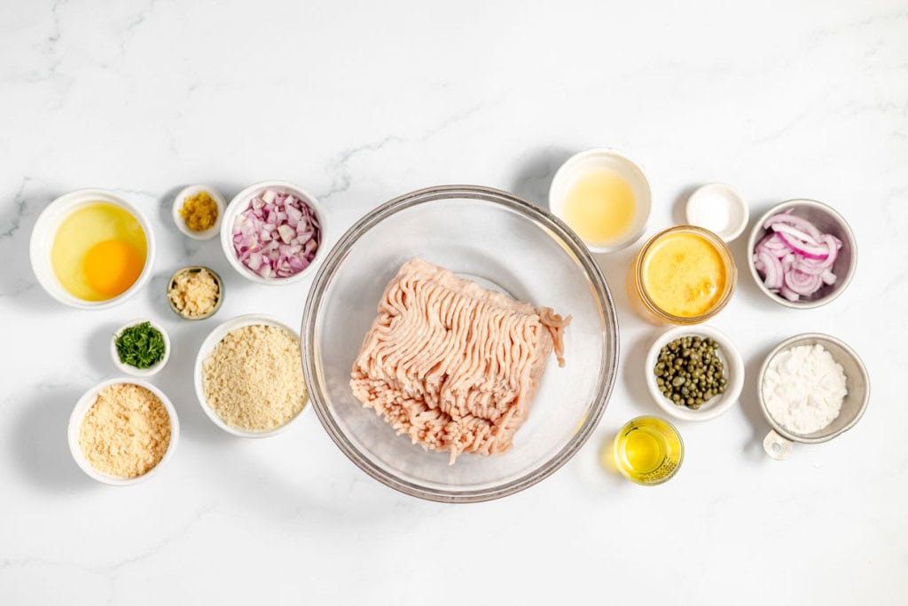 A bowl of ground turkey surrounded by various ingredients in small bowls, including eggs, onions, breadcrumbs, spices, capers, oil, and lemon juice on a white marble surface.