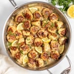 A skillet with cooked browned meatballs in a lemon herb sauce, garnished with chopped parsley and lemon wedges. A white cloth and a cut lemon are placed beside the skillet on a white surface.