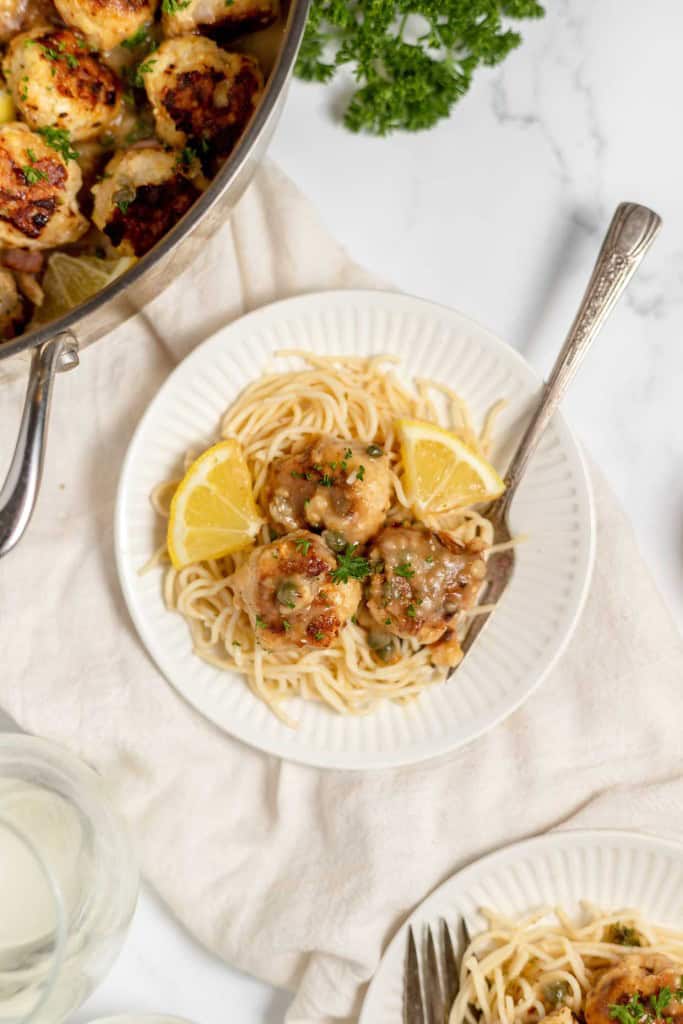 A plate of spaghetti topped with meatballs garnished with lemon wedges and parsley, with a fork on the side. A pan with more meatballs and a sprig of parsley are visible in the background.