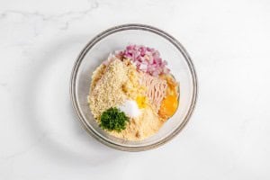 A glass bowl containing minced meat, breadcrumbs, chopped onions, one egg, chopped parsley, salt, and grated cheese on a marble countertop.
