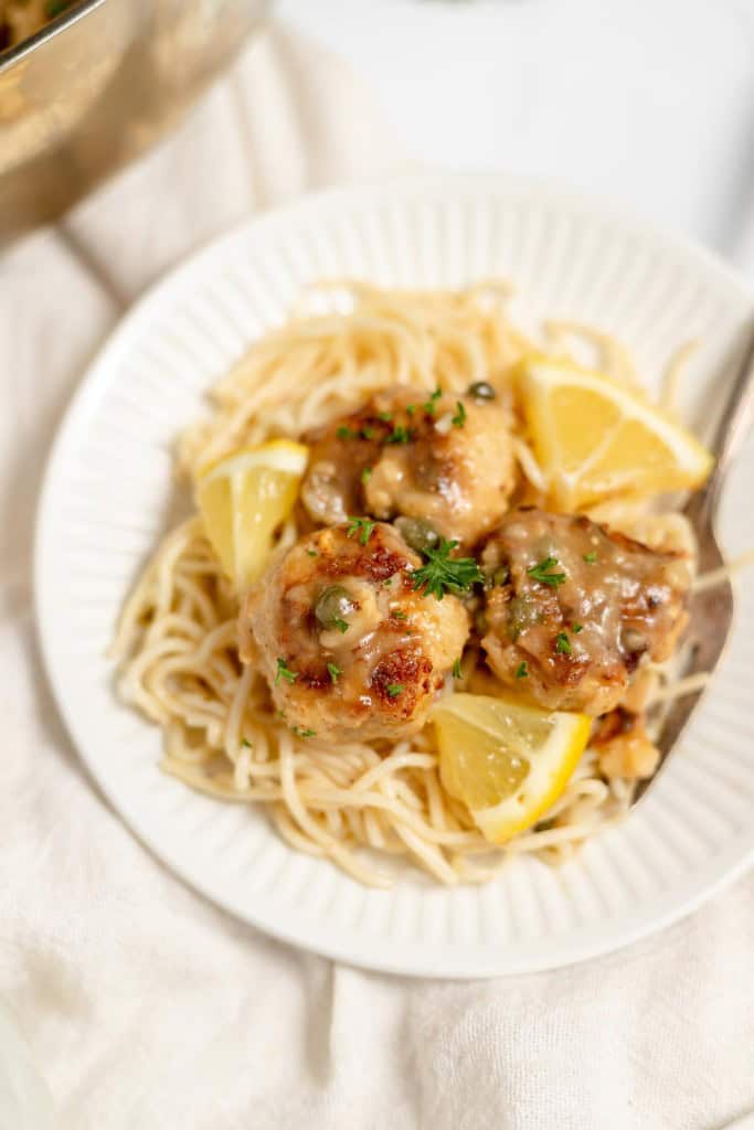 A white plate with spaghetti topped with meatballs in a creamy sauce, garnished with chopped parsley and lemon wedges.