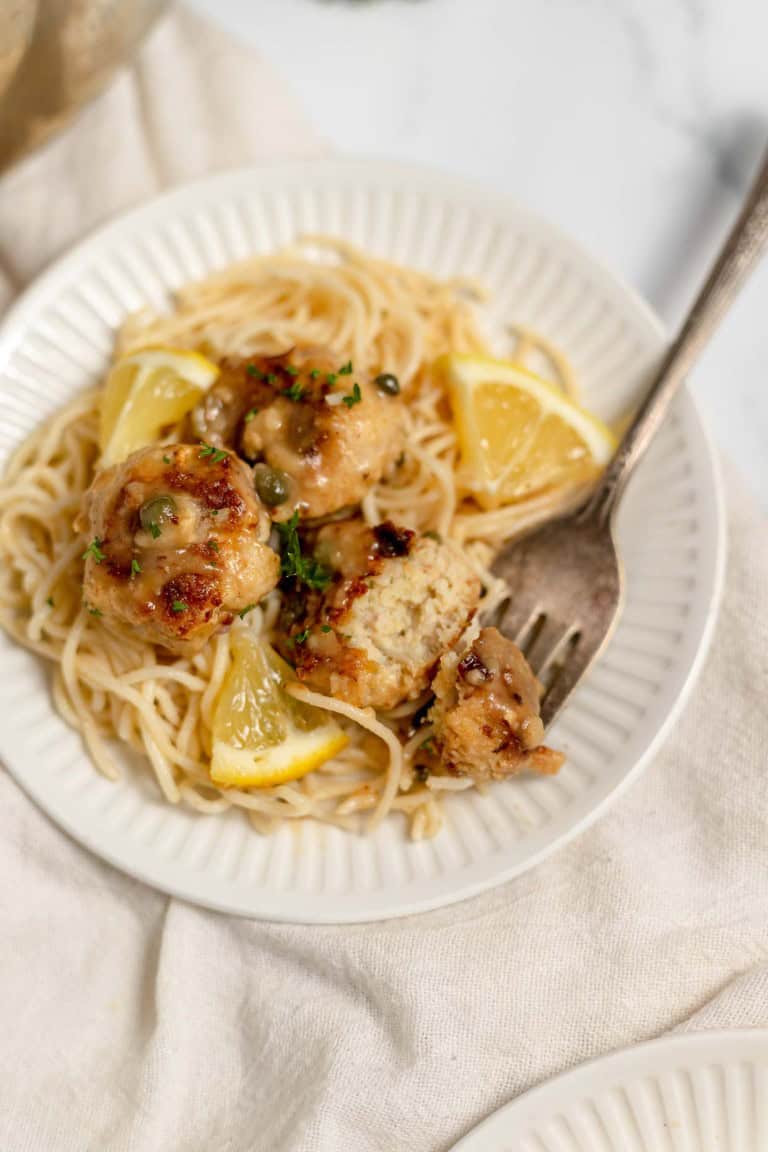 A white plate with spaghetti, lemon wedges, and chicken meatballs garnished with capers and parsley. A fork rests on the plate.