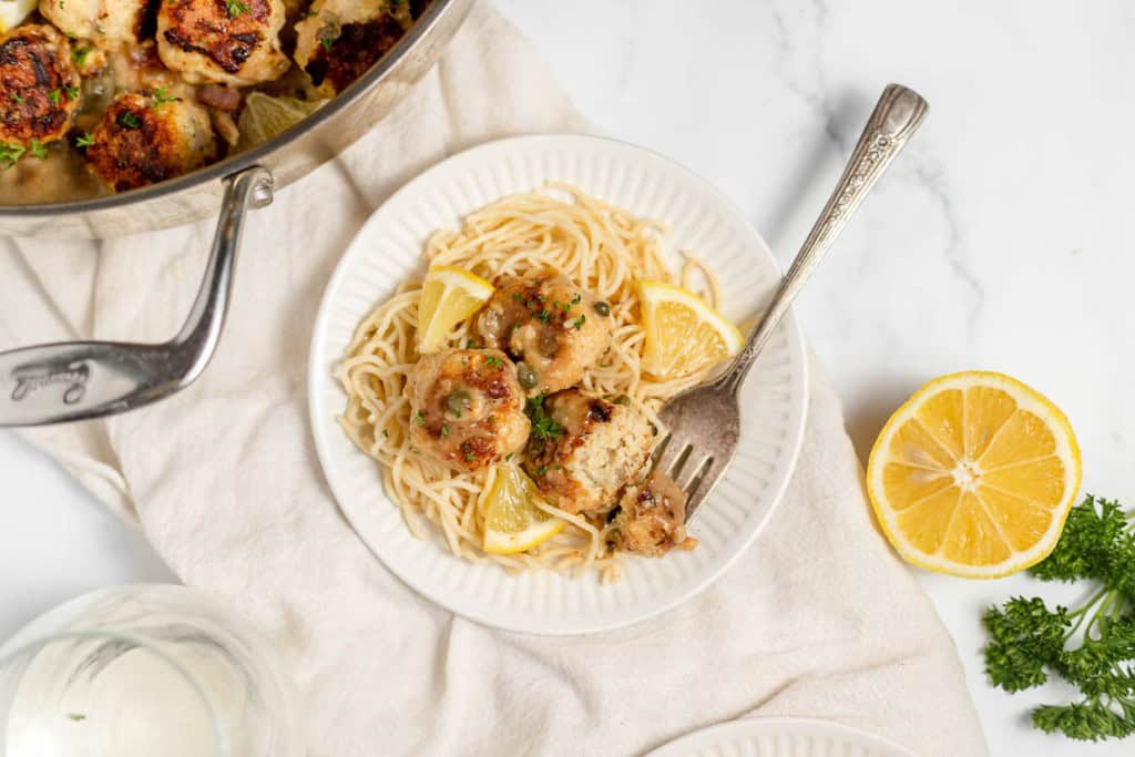 A plate of pasta with meatballs garnished with lemon slices and parsley. A pan with more meatballs is visible next to the plate. A fork and half a lemon are on the side.