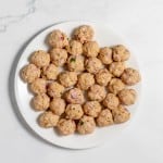 A white plate holds numerous uncooked meatballs, mixed with diced onions and herbs, arranged in a circular pattern on a white marbled surface.