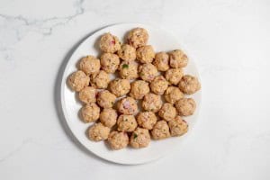 A white plate holds numerous uncooked meatballs, mixed with diced onions and herbs, arranged in a circular pattern on a white marbled surface.