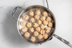A frying pan containing evenly spaced, partially cooked meatballs on a marble countertop.
