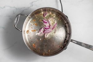 A stainless steel pan on a marble surface contains sautéing sliced red onions with olive oil and browned residue on the bottom.