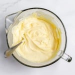 A glass mixing bowl filled with creamy yellow batter, hinting at a luscious lemonade pie, with a metal spoon sticking out from the side, placed on a white marble countertop.