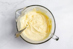 A glass mixing bowl filled with creamy yellow batter, hinting at a luscious lemonade pie, with a metal spoon sticking out from the side, placed on a white marble countertop.