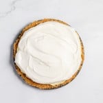A lemonade pie with a graham cracker crust and a topping of whipped cream rests on a white marble surface.