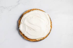A lemonade pie with a graham cracker crust and a topping of whipped cream rests on a white marble surface.