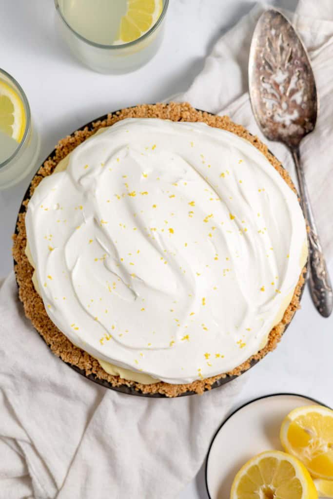 A lemonade pie topped with whipped cream and lemon zest, accompanied by two glasses of lemonade and lemon slices, on a white cloth with a vintage metal serving spoon nearby.