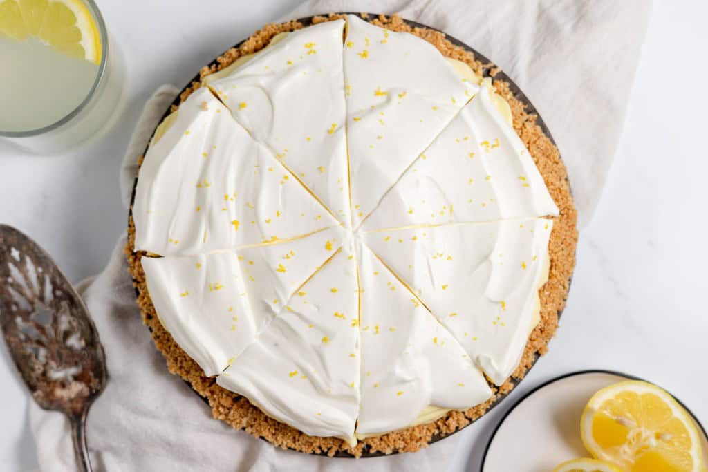 A sliced lemonade pie with a graham cracker crust, topped with whipped cream and lemon zest. A nearby slice is plated alongside lemon slices and a glass of lemon water.