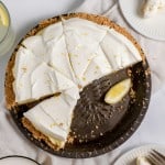 A lemonade pie with a slice missing, topped with lemon zest, sits on a dark pie dish. Two plates with pie slices, one adorned with a lemon wedge, and two glasses of refreshing lemonade are artfully arranged around it.