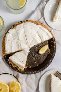 A lemonade pie with a slice missing, topped with lemon zest, sits on a dark pie dish. Two plates with pie slices, one adorned with a lemon wedge, and two glasses of refreshing lemonade are artfully arranged around it.