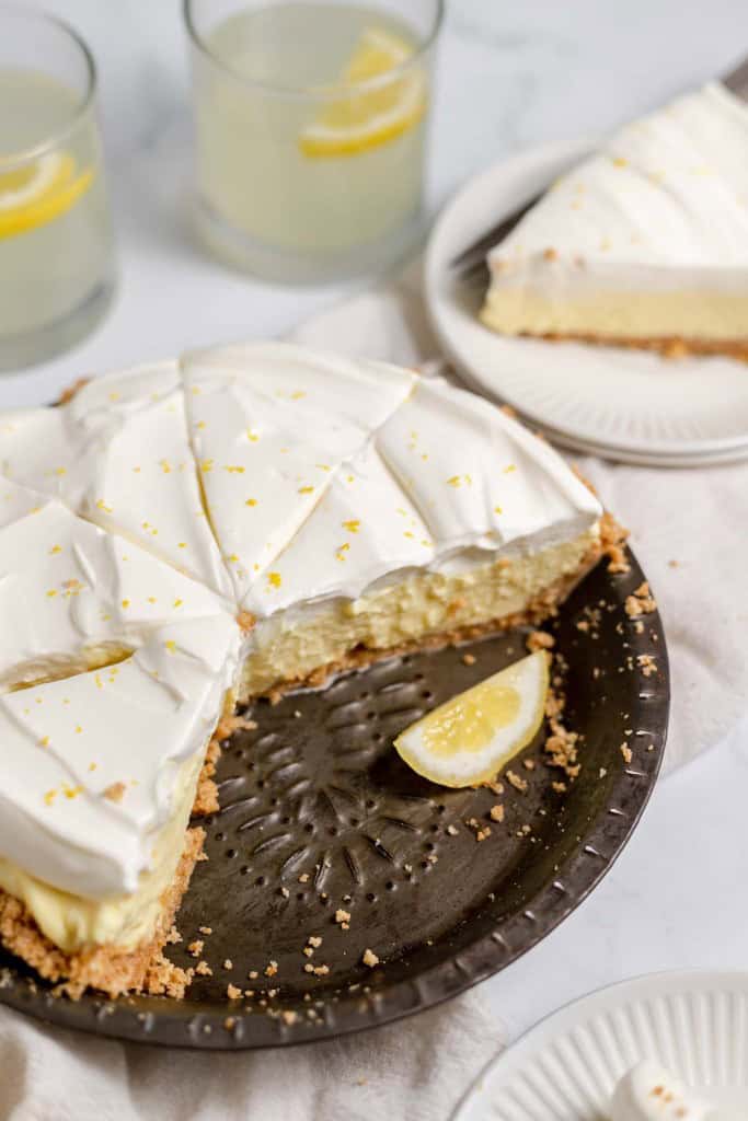 A lemonade pie with a creamy white topping in a metal pie dish, missing one slice. A slice of the pie is on a plate in the background, near two glasses filled with a light yellow beverage, each containing a lemon wedge.