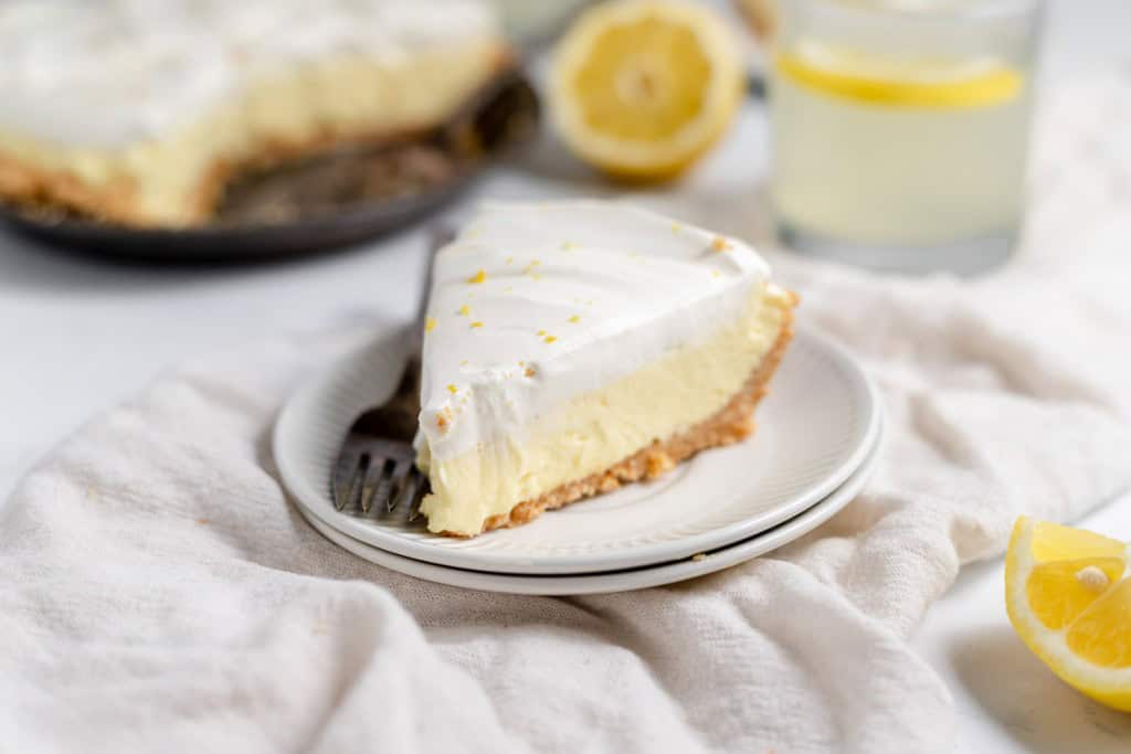 A slice of lemonade pie with a graham cracker crust sits on a white plate, accompanied by a fork. A glass of lemon water and additional pie are in the background.