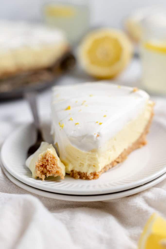 A slice of lemonade pie on a white plate with a fork. The pie has a creamy filling, topped with whipped cream and lemon zest. Blurred lemon and pie in the background.