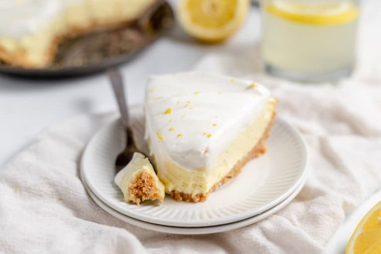 A slice of lemonade pie on a white plate with a fork holding a small piece. A whole pie and a glass of refreshing lemon drink are in the background.