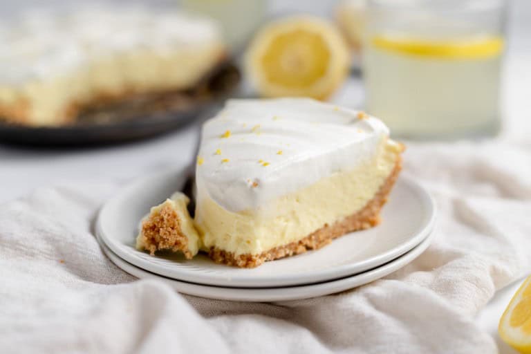 A slice of lemonade pie with a graham cracker crust and whipped topping sits on a white plate, with a glass of lemon water and a lemon half in the background.