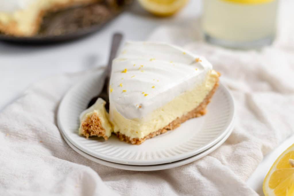 A slice of lemon cheesecake with a graham cracker crust is on a white plate with a forkful beside it. A refreshing glass of lemonade pie cocktail and a lemon are in the background, adding to the zesty delight.