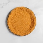 A round, partially baked lemonade pie crust with a golden-brown crumb texture sits on a light marble surface.