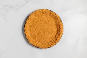 A round, partially baked lemonade pie crust with a golden-brown crumb texture sits on a light marble surface.