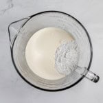 A glass measuring cup containing a mixture of creamy white liquid and white powder, reminiscent of the beginnings of a classic lemonade pie, sits against a white marble background.