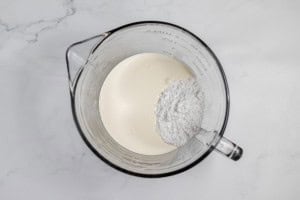 A glass measuring cup containing a mixture of creamy white liquid and white powder, reminiscent of the beginnings of a classic lemonade pie, sits against a white marble background.
