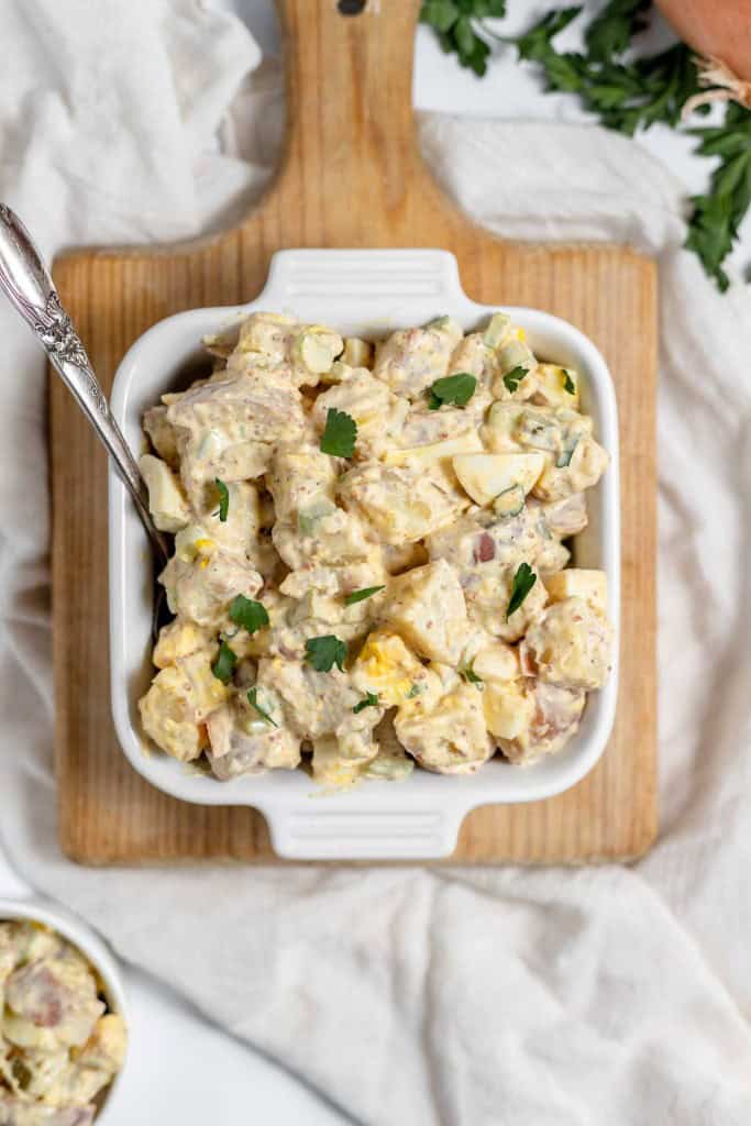 A bowl of creamy mustard potato salad with eggs and herbs on a wooden board, garnished with parsley, against a white cloth background.