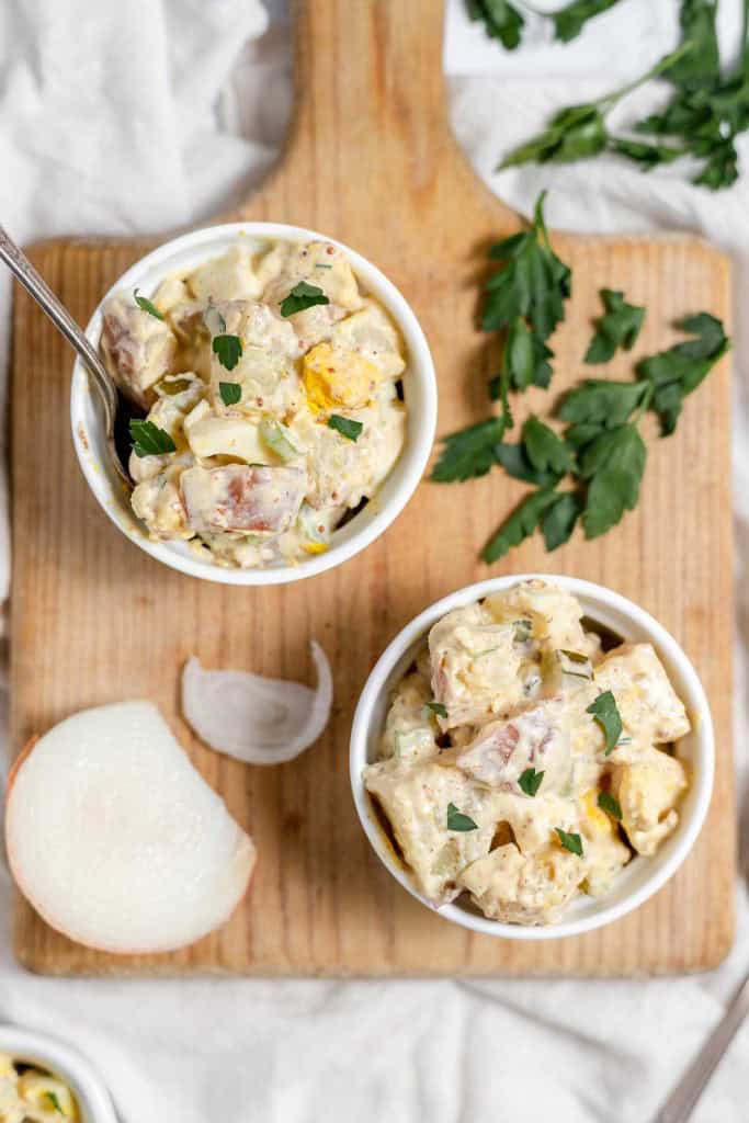 A bowl of mustard potato salad with eggs and herbs on a wooden cutting board, next to raw onion and garlic cloves.