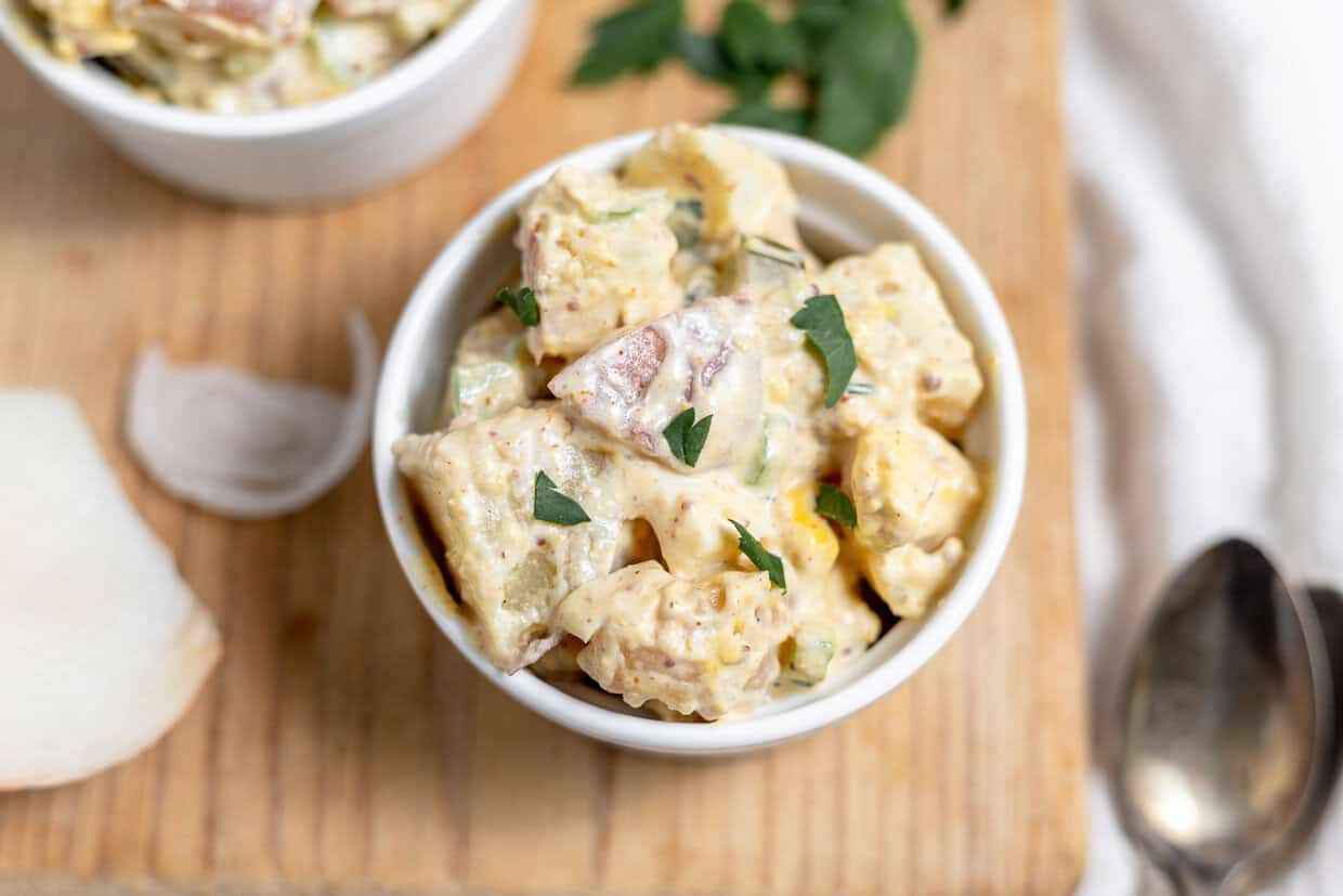 A bowl of creamy mustard potato salad garnished with parsley, served on a wooden table with a spoon and ingredients in the background.
