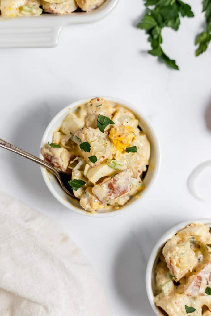 A bowl of creamy mustard potato salad garnished with parsley, served on a white background with a spoon.