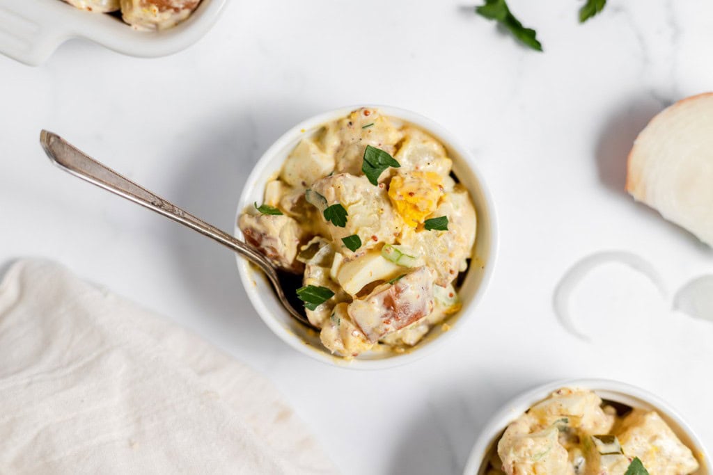 A bowl of mustard potato salad garnished with herbs, with a spoon, on a white marble surface next to sliced onions.