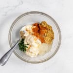 A bowl containing ingredients for mustard potato salad including spices, herbs, and condiments, viewed from above on a marble surface.