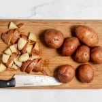 Several whole and chopped red potatoes on a wooden cutting board alongside a kitchen knife, set on a marble countertop for making mustard potato salad.