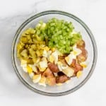 Top view of a glass bowl with a salad containing diced sausages, boiled eggs, green peppers, mustard potato salad, and pickles on a marble surface.