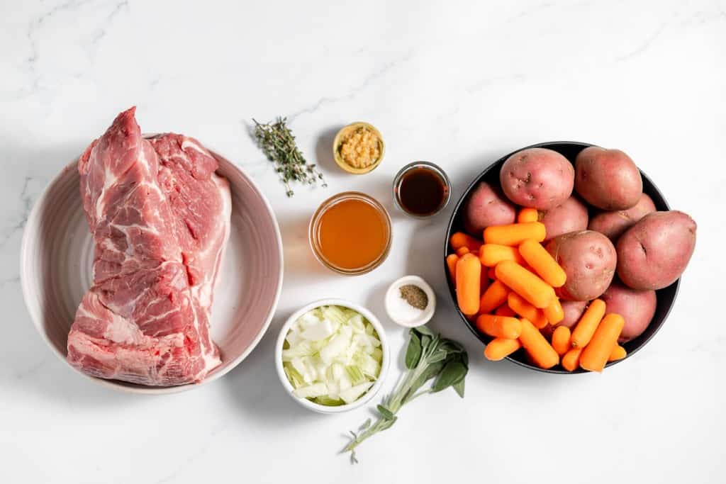 Raw pork on a plate, with bowls of carrots, potatoes, onions, and sauces on a marble surface.