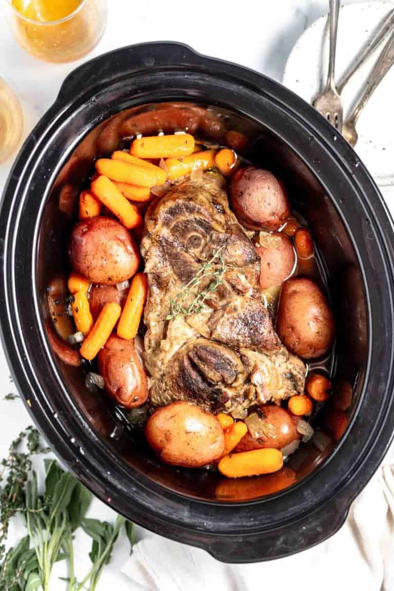A slow cooker filled with a cooked lamb shank, carrots, and potatoes, surrounded by herbs on a white surface.
