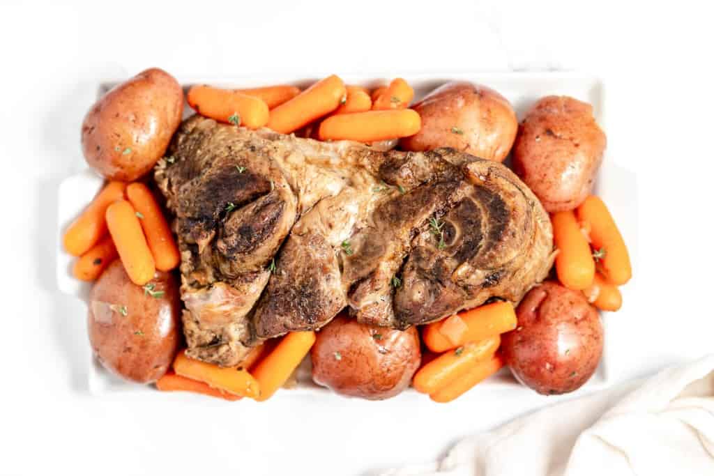 Roasted lamb shanks on a white platter with baby carrots and potatoes, shot from above.