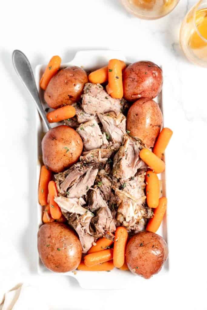 A platter of roast pork surrounded by carrots and potatoes, served with a spoon on a white marble countertop.
