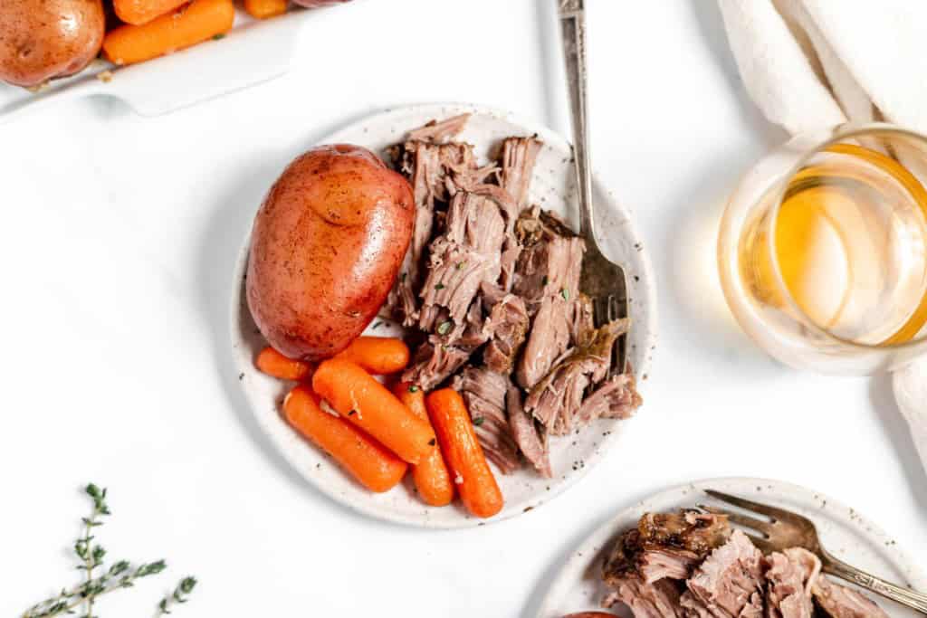 A plate of pulled beef with whole potatoes, baby carrots and a glass of juice on a white tabletop.
