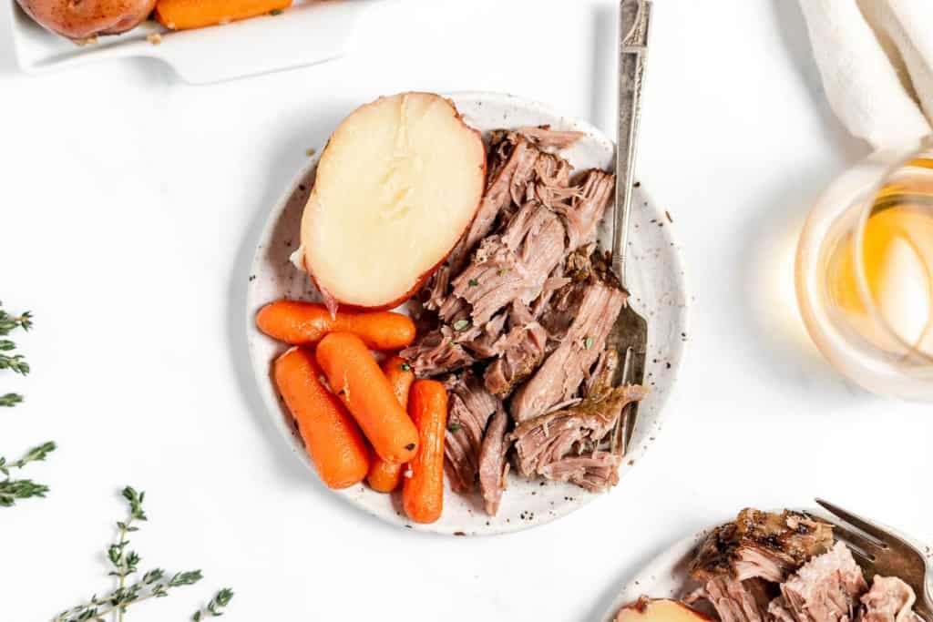 Sliced pot roast with carrots and a halved potato on a speckled plate, with a glass of cider and herbs nearby, on a white background.
