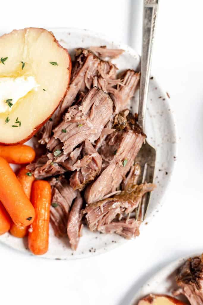 A plate of pulled pork with carrots and a baked potato topped with butter, garnished with herbs.